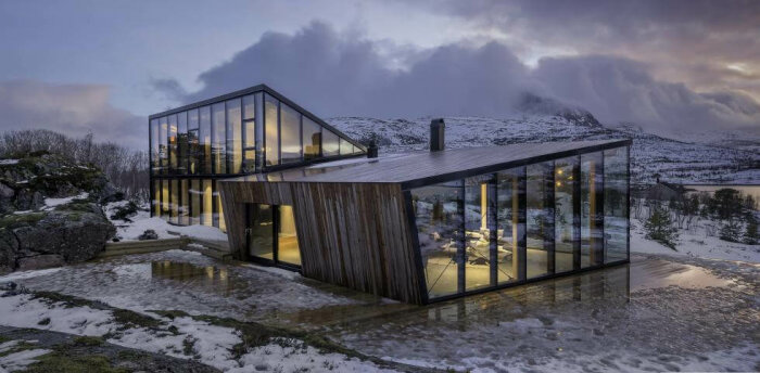 Modernt hus med glasfasader i vacker, snöig fjällmiljö vid skymning, med reflekterande ljus i vattenpölar.