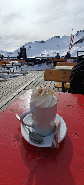 En kopp varm dryck med vispgrädde på ett rött bord utomhus, omgiven av snötäckta berg.