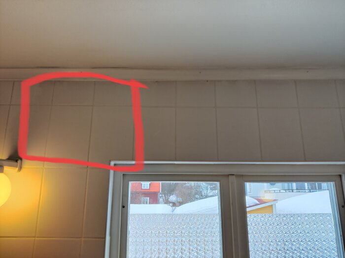 Inomhusvy med fönster, frostad glasruta, snö utsikt, markerad plats med röd cirkel, väggbelysning, kakelvägg.