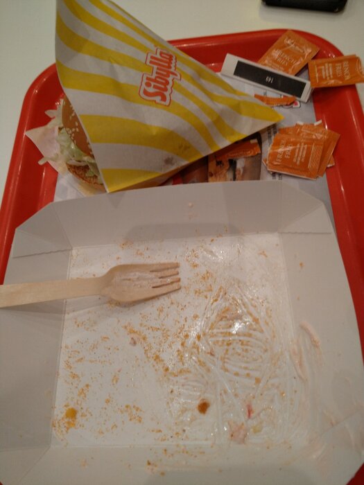 Ett matfat efter måltid med smulor, tom hamburgerförpackning, såspåsar och träbestick på röd bricka.