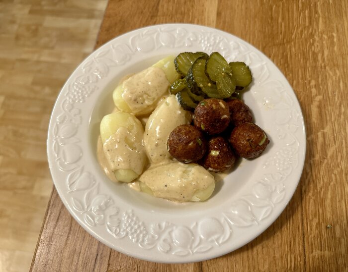 Tallrik med köttbullar, potatis, gräddsås och skivade inlagda gurkor, på träbord.