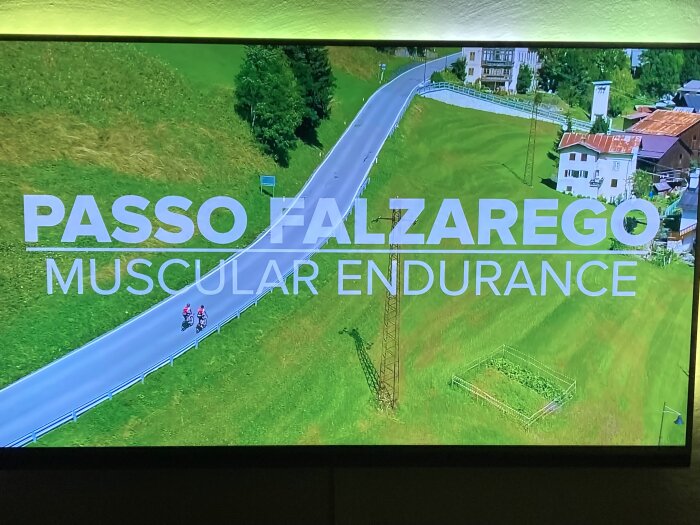 Luftfoto av landsväg, text "PASSO FALZAREGO MUSCULAR ENDURANCE", cyklister, gröna fält, byggnader.