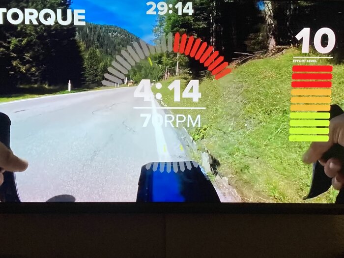 Indoor cycling skärm som visar träning, vägläge, motstånd, kadens och ansträngningsnivå mot gröna skogsbakgrunder.