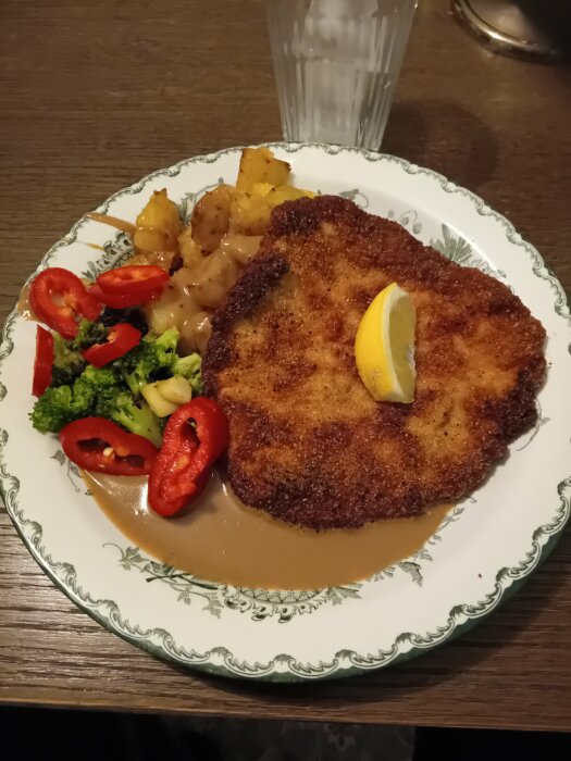 En tallrik med schnitzel, sås, citronskiva, stekt potatis, broccoli och röd paprika.