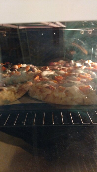 Hemlagade pizzor gräddas i en ugn; synliga genom glaslucka med ånga och reflektioner.