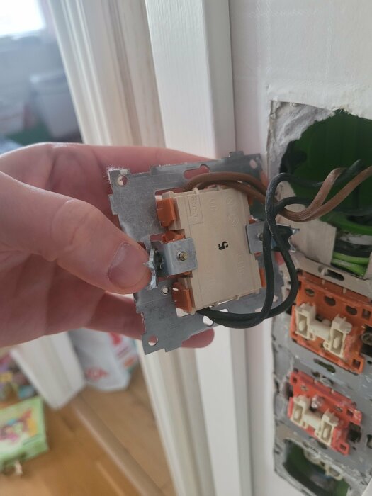 En hand håller en delvis demonterad eluttag framför en öppen vägg med synliga kablar och andra eluttag.