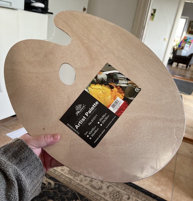 En person håller en ny och oanvänd konstnärs palett i trä, fortfarande i plastförpackning.