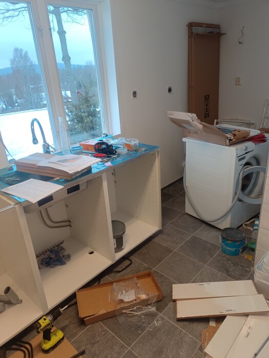 Kök under renovering, osammanbyggda skåp, verktyg, rörigt, snö utanför fönstret.