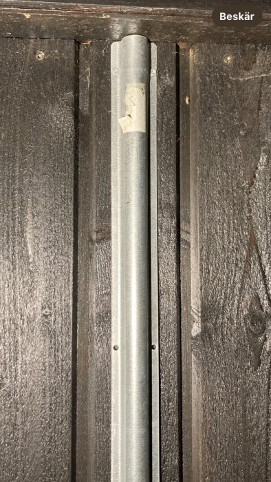 Metallrör mot mörkt trä. Vertikal position. Skruvar synliga. Slitage tecken. Enkel, industriell estetik. Beskär text övre högra hörnet.