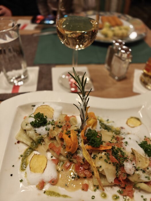 Gourmeträtt med pasta, skum, krispiga chips, persilja, tomat, i elegant restaurangmiljö med vin och vattenglas.