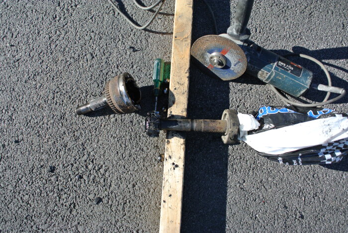 Verktyg och bilkomponenter på asfalt: domkraft, mejsel, slipskiva, grus, trasig bilaxel med hjulnav.