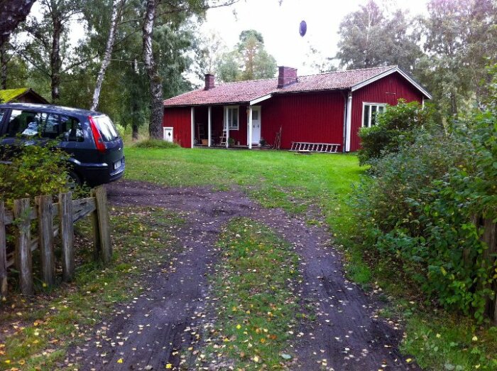 Rött äldre hus med vita knutar och källardörr syns bakom en grön gräsmatta och en grusväg med en parkerad bil till vänster.