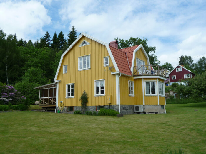 Gult trähus med vita knutar, röd tak, grönt gräs, träd och altan, klassisk svensk stil.