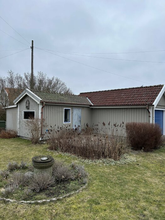 Ett litet hus med tegeltak, gråa väggar, fönsterluckor och en vissnad trädgård.