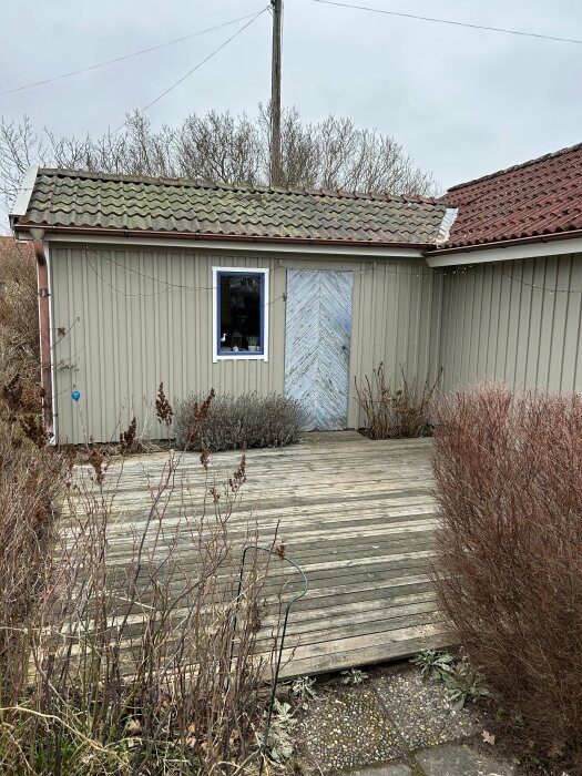 En småsliten trädäck leder till ett brunt hus med slitna takplattor och en blå dörr.