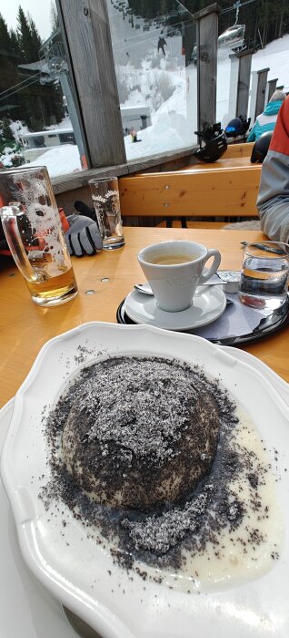 Dessert-dumpling med pudrat socker, omgiven av vaniljsås, på ett bord framför ett kaffekopp och ölglas, med skidbacke i bakgrunden.