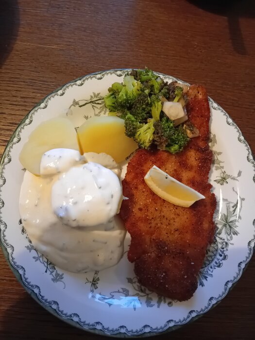 Panerad flundra med en klick gräslöksmajonnäs, kokta potatisar och ångad broccoli på ett mönstrat fat.