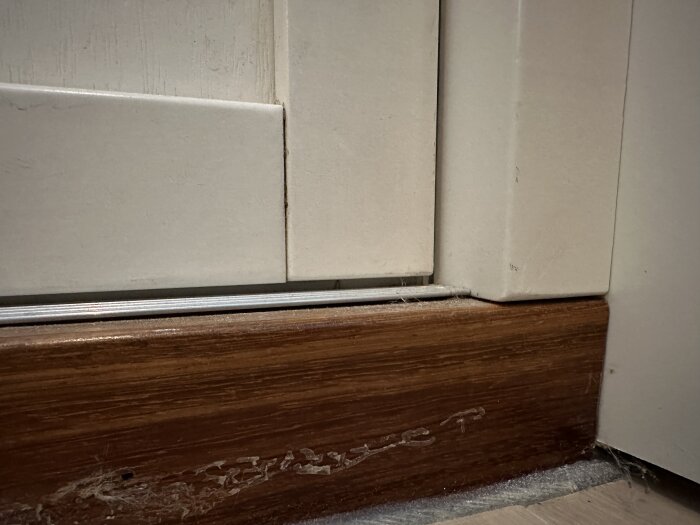 Närbild av rödbrun fotlist vid vit vägg och kaklat golv med synlig slitage och rester av byggmaterial.