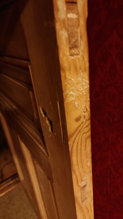 Närbild på en träpanel med repor och skador längs kanten mot en röd mönstrad vägg.