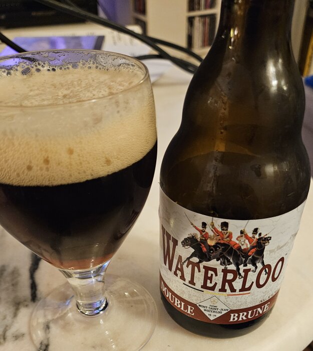 Ett glas med mörkt öl och en öppnad flaska Waterloo Double Brune på ett bord.