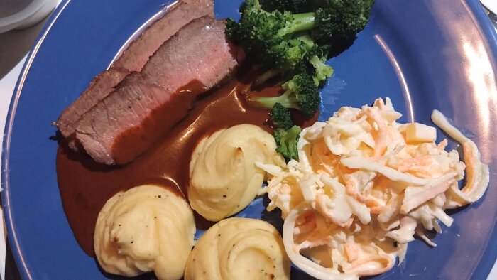 Ett måltid med kött, potatisduchesse, broccolibuketter, sås och coleslaw på en blå tallrik.