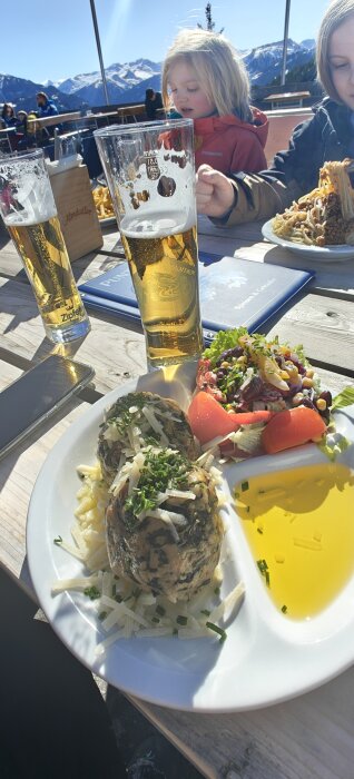 Spinatknödel på tallrik med sallad, riven ost och ölglas på ett bord med bergsutsikt.