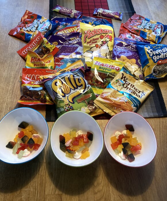 Tre skålar med blandat godis framför en samling godispåsar på ett bord.