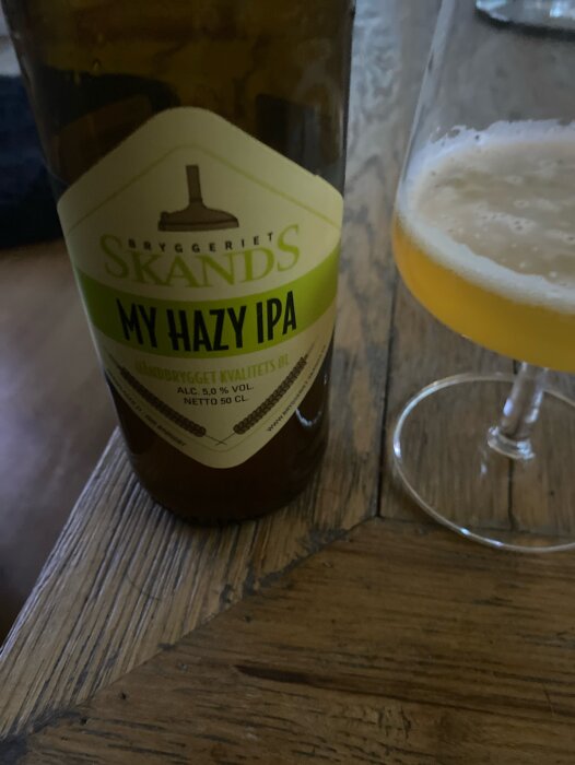 Flaska av Skands Bryggeriet My Hazy IPA med synlig etikett bredvid ett ölglas med bubblig IPA på ett träbord.