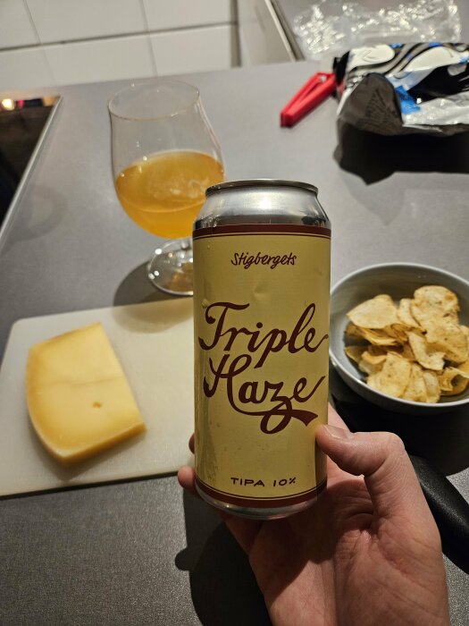 Hand håller en ölburk med texten "Triple Haze TIPA 10%", glas med öl och ostbit i bakgrunden.