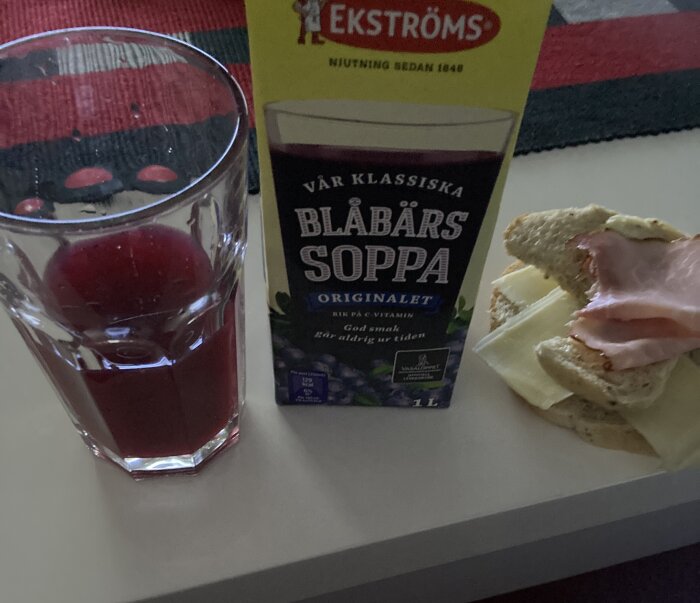 Ett glas med röd dryck, förpackning av blåbärssoppa och en smörgås med ost och skinka.