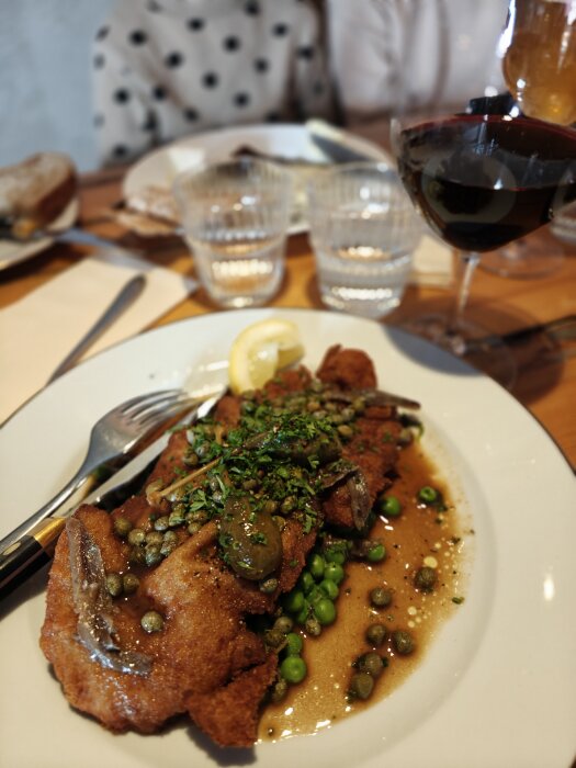 Schnitzel med kapers, ärtor och champinjonsås på en tallrik, med ett glas rödvin i bakgrunden.