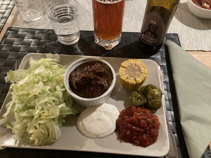 Serverad texasgryta med strimlad sallad, gräddfil, salsa, jalapeños och majs på en vit bricka med öl.