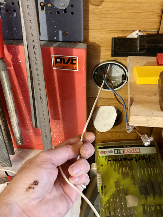 Hand håller en kabel nära en öppen vägguttag och ett verktygsfodral i en verkstadsinställning.