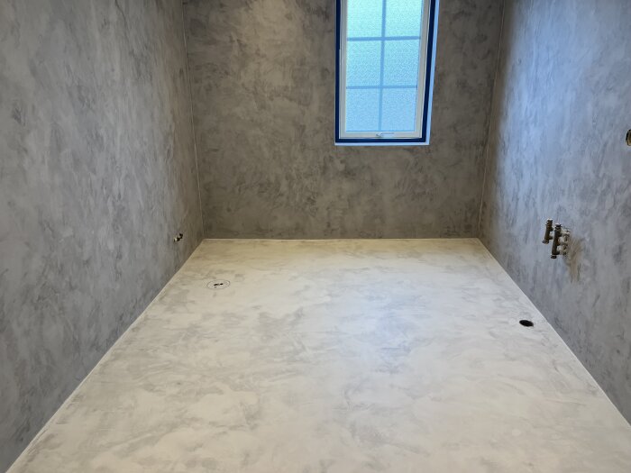 Badrum under renovering med marmorliknande microcement på väggar och golv, blått fönster och rörinstallationer.