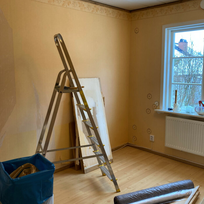 Renovering pågår i tomt rum med stege, gipsplattor, tapetrulle och skyddsplast på golvet.