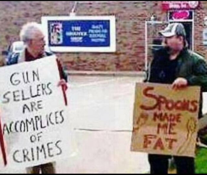 Två personer håller protestskyltar, en med texten "GUN SELLERS ARE ACCOMPLICES OF CRIMES", en med "SPOONS MADE ME FAT".