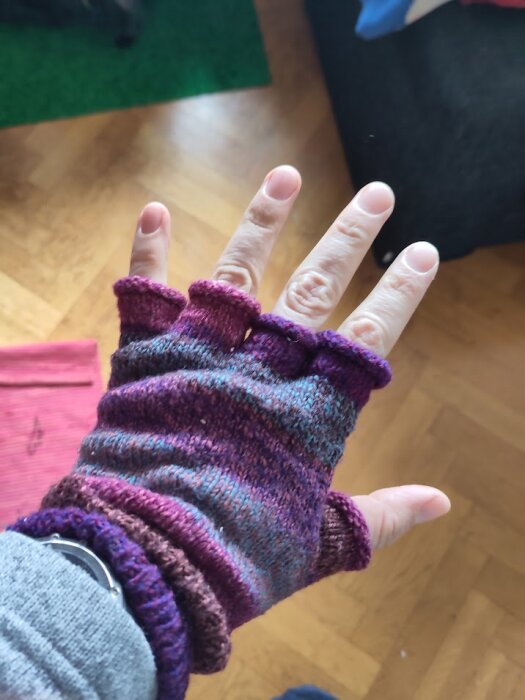 Hand i fingerlösa vantar i nyanser av lila och blått mot en bakgrund av trägolv.