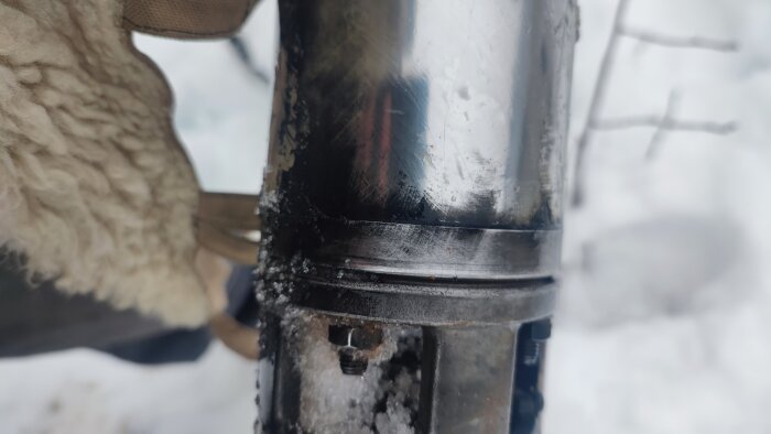 Närbild av en svart föremål med frost, snöig bakgrund, detaljer suddiga, delvis synlig vintervante.