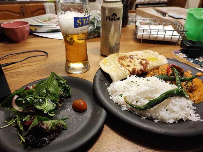 Middag med ris, paneer, sallad, öl och chilipeppar på ett bord. Oorganiserad bakgrund, vardaglig inomhusmiljö.