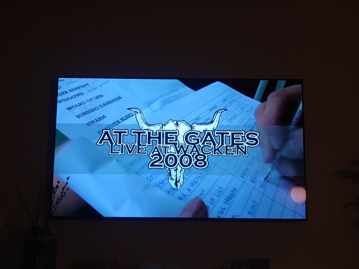 TV-skärm som visar texten "AT THE GATES LIVE AT WACKEN 2008" ovanför en hand som skriver på ett papper.
