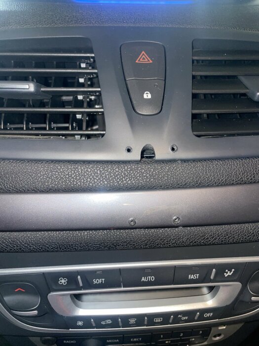 Bilens mittkonsol med A/C-ventiler och kontrollknappar, två synliga skruvhål under en varningstriangelknapp.
