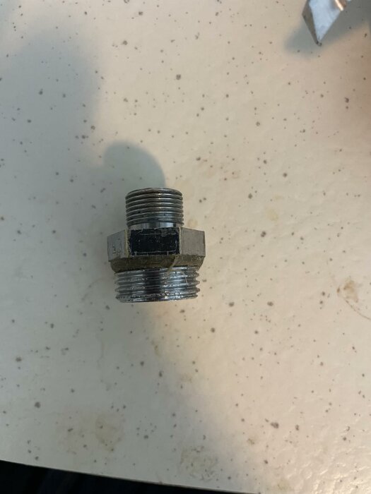 Metallisk anslutningsnippel med gängor, ligger på en ljus yta med små fläckar.