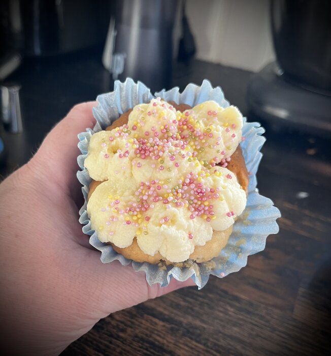 Ett muffinsformat bakverk med krämig topping och färgglada strössel, hålls i handen.