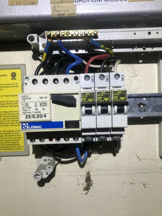 Elcentral med säkringar, jordfelsbrytare och ledningar på en vägg. Tejpade etiketter och testdatum synliga.