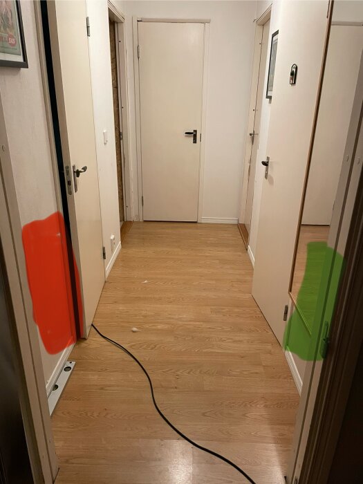 Korridor med trägolv och flera dörrar, markerad med en röd färg där golvvärmeskåpet ska placeras, och en grön färg vid ventilationsschaktet.