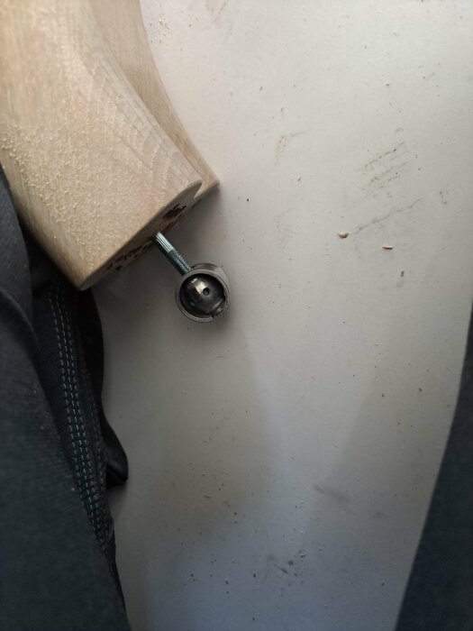 Trädel med utstickande skruv, nära en skräpig, grå yta. Del av svart textil synlig.