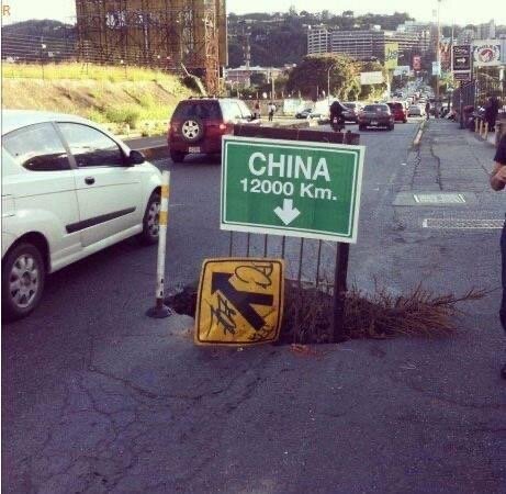 Skylt som visar "CHINA 12000 Km" med ikon för arbetsområde, på sprucken väg med bilar i bakgrunden.