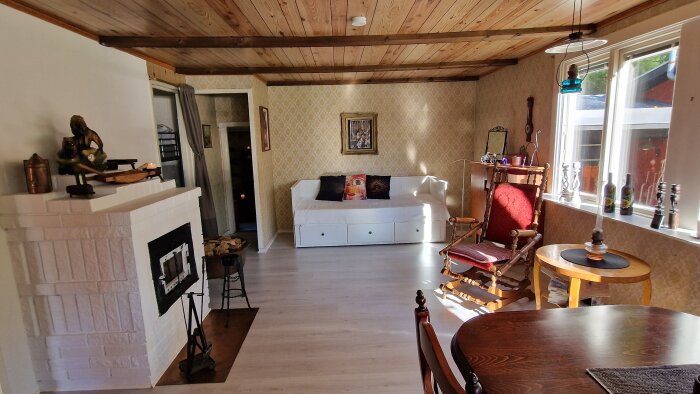 Ett ljust vardagsrum med vedspis, soffa, trägolv, takbjälkar och vintage inredning. Soligt fönster till höger.