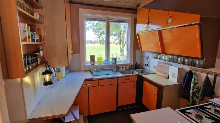 Retrokök med orange skåpluckor, diskho, fönster med utsikt och kryddhylla. Lantlig stil, dagsljus.