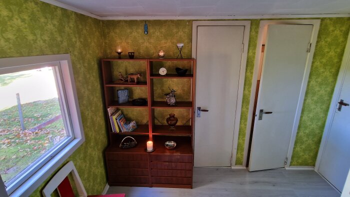 Rum med grön tapet, trähylla, dekorationer, spegel, ljus, dörrar och ett fönster med utsikt mot trädgård.